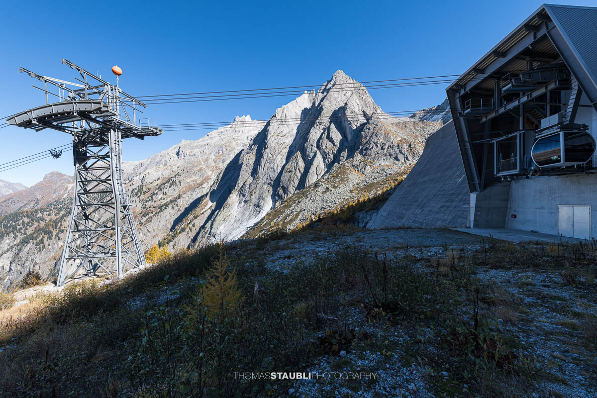Die Bergstation der Luftseilbahn am Albignasee steht am Rand eines alpinen Plateaus, flankiert von einem markanten Stahlmast mit darüber verlaufenden Seilen. Dahinter ragen steile, graue Granitflanken und ein scharf geschnittener Gipfel in den klaren Herbsthimmel, während am Hang goldene Lärchen leuchten.