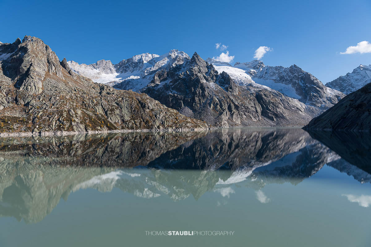 Klare Spiegelung der schroffen Bergeller Alpen im türkisgrünen Wasser des Albignasees, umgeben von Felsflanken und Gletschern unter einem wolkenlosen blauen Himmel.