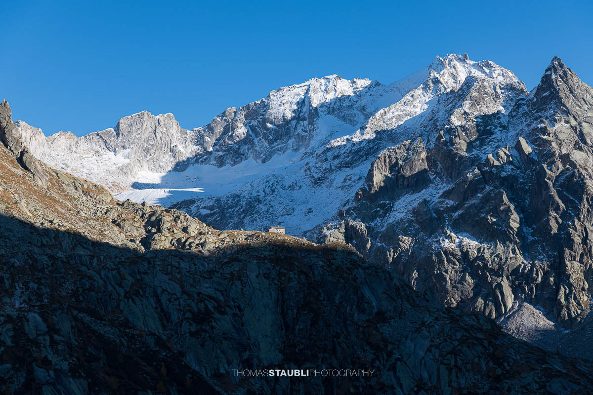 Die Albignahütte liegt im warmen Licht der Abendsonne auf einem Felsvorsprung, dahinter erheben sich die schroffen Gipfel und der Gletscher Vadrec dal Cantun in den Bergeller Alpen.