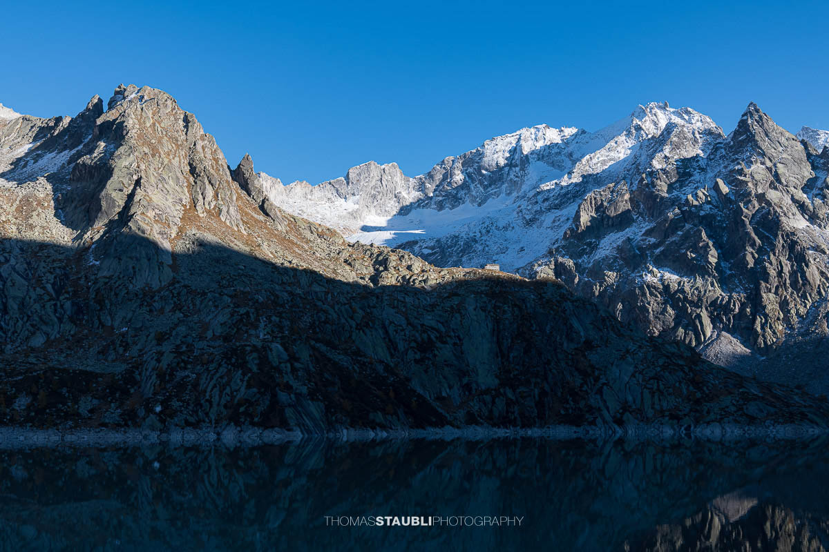 Die Albignahütte liegt im warmen Licht der Abendsonne auf einem Felsvorsprung, dahinter erheben sich die schroffen Gipfel und der Gletscher Vadrec dal Cantun in den Bergeller Alpen.
