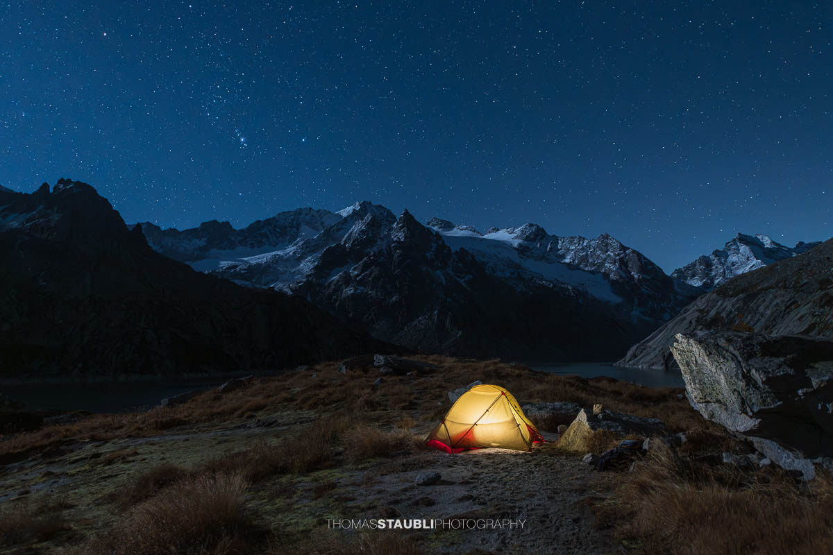 Ein beleuchtetes Zelt steht unter einem klaren Sternenhimmel am Ufer des Albignasees im Bergell. Im Hintergrund erheben sich die schneebedeckten Gipfel und Gletscher der Bergeller Alpen, während das sanfte Licht des Zelts Wärme in die kühle Bergnacht bringt.