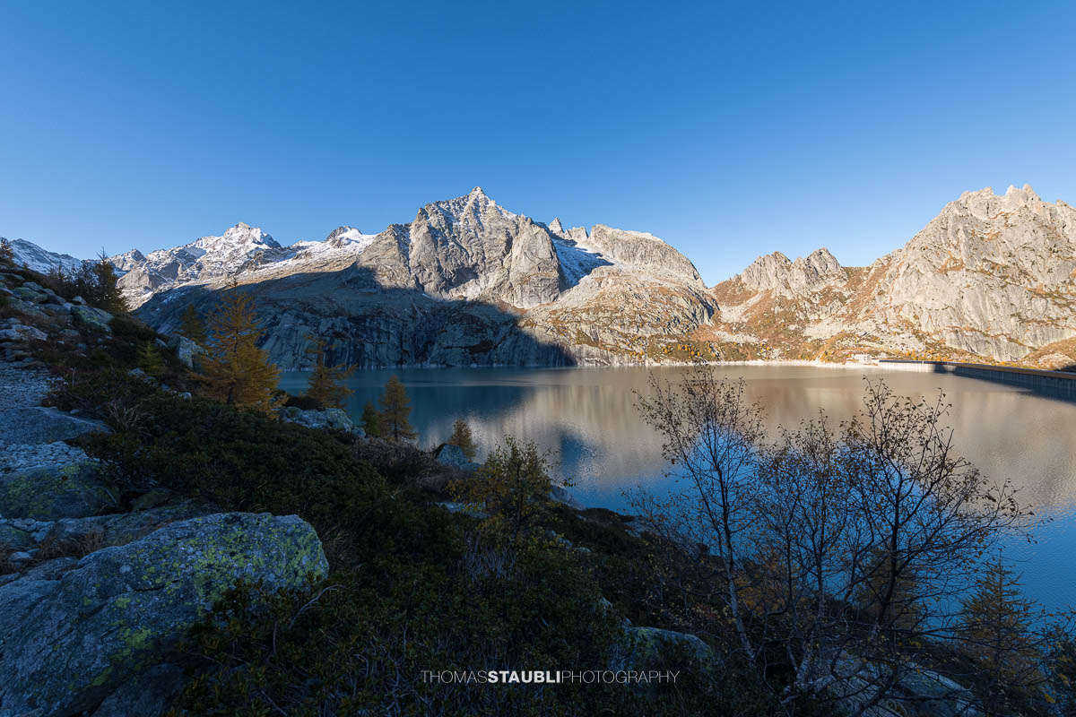 Klare Herbstluft und frühes Sonnenlicht tauchen den Albignasee im Bergell in kühle Farben. Die umliegenden Granitgipfel spiegeln sich im stillen Wasser, während die ersten Lärchen am Ufer golden leuchten. Im Hintergrund ragen die verschneiten Berge der Bergeller Alpen auf.