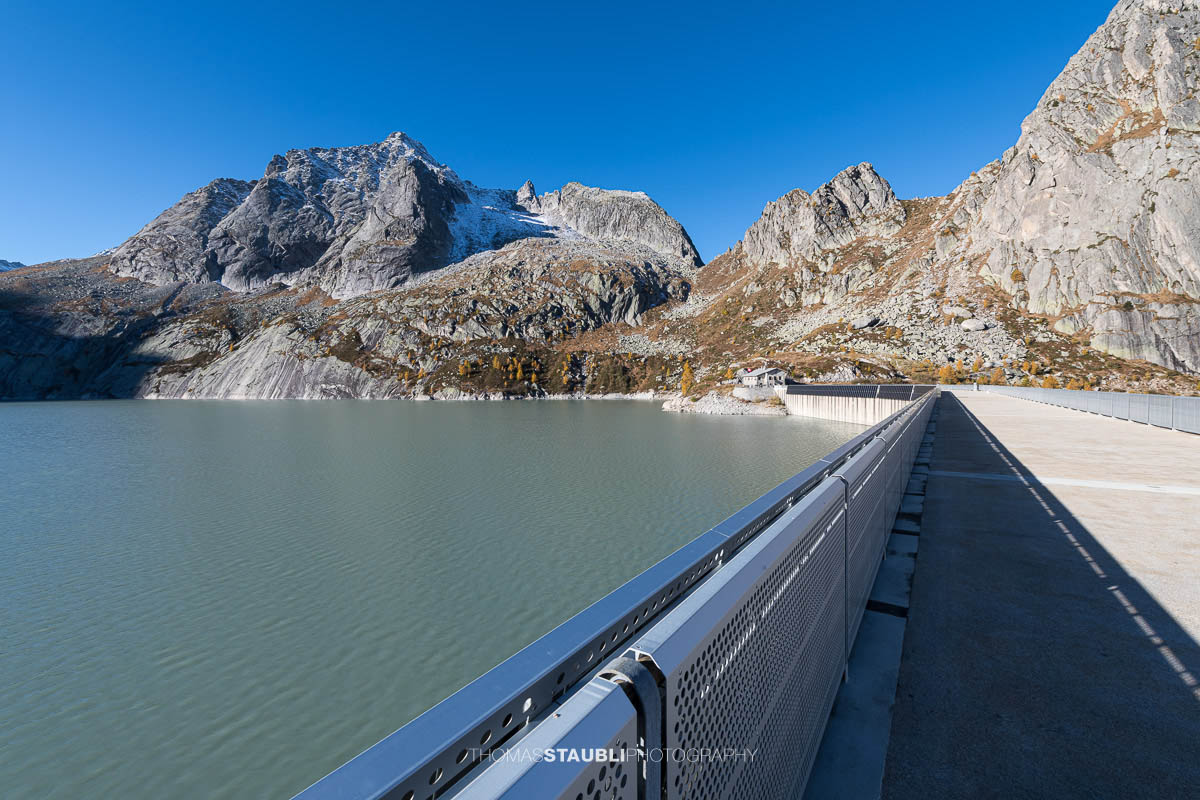 Blick über die Staumauer des Albignasees im Bergell. Im klaren Morgenlicht kontrastiert die moderne Metallkonstruktion mit der rauen Granitlandschaft. Auf der gegenüberliegenden Seite liegt das Betriebsgebäude der Albigna-Staumauer, umgeben von den steilen, teils verschneiten Gipfeln der Bergeller Alpen.