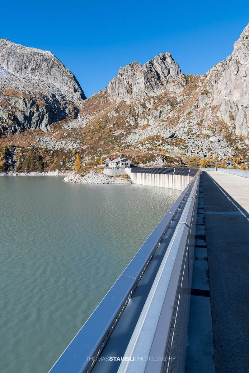 Blick über die Staumauer des Albignasees im Bergell. Im klaren Morgenlicht kontrastiert die moderne Metallkonstruktion mit der rauen Granitlandschaft. Auf der gegenüberliegenden Seite liegt das Betriebsgebäude der Albigna-Staumauer, umgeben von den steilen, teils verschneiten Gipfeln der Bergeller Alpen.
