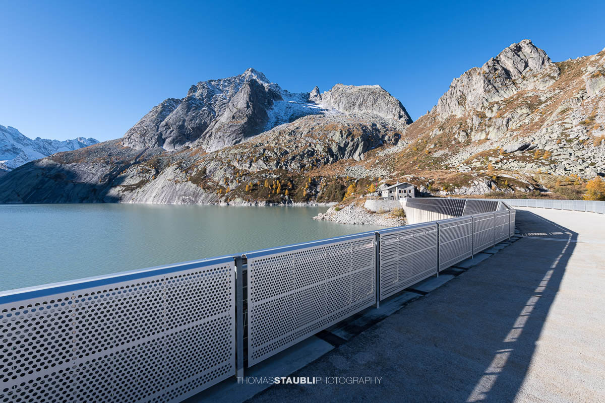 Blick über die Staumauer des Albignasees im Bergell. Im klaren Morgenlicht kontrastiert die moderne Metallkonstruktion mit der rauen Granitlandschaft. Auf der gegenüberliegenden Seite liegt das Betriebsgebäude der Albigna-Staumauer, umgeben von den steilen, teils verschneiten Gipfeln der Bergeller Alpen.