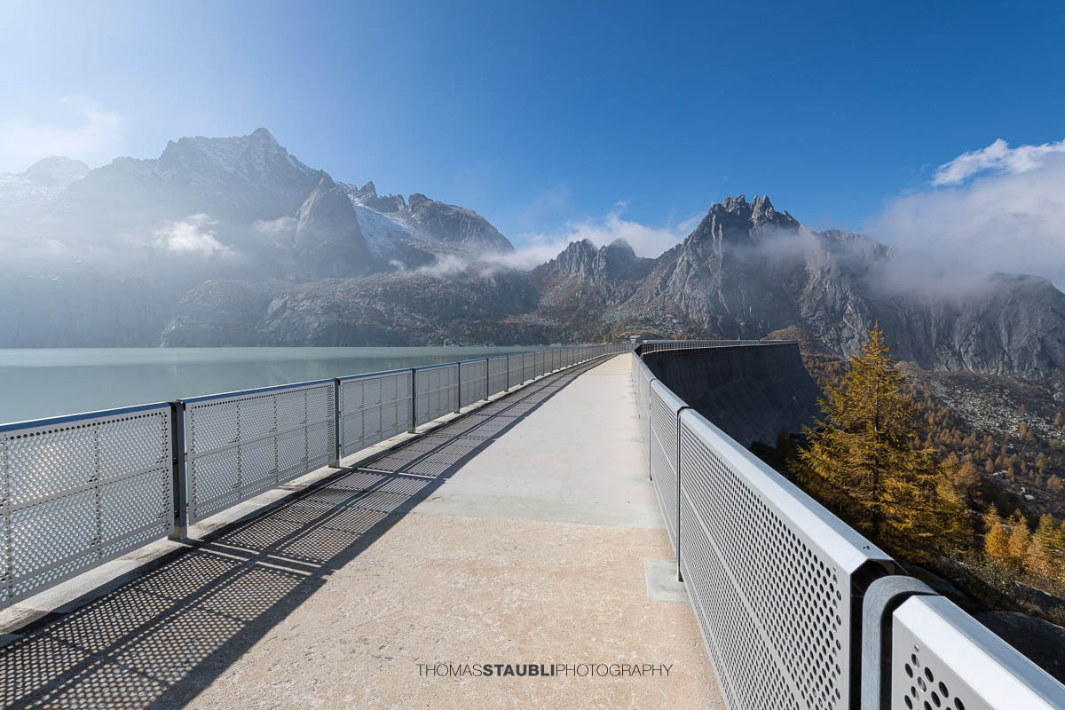 Blick über die begehbare Staumauer des Albignasees, die als schmaler Weg zwischen Metallgeländern verläuft. Links liegt der ruhige, milchig-türkise See, rechts fällt die Mauer steil ab. Im Hintergrund ragen schroffe Granitgipfel mit leichtem Schnee und vorbeiziehenden Nebelschwaden in den klaren blauen Himmel.
