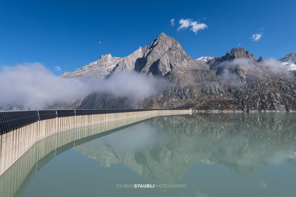 Die geschwungene Staumauer des Albignasees spiegelt sich im milchig-türkisfarbenen Wasser, während über ihr einzelne Wolkenfetzen hängen. Dahinter ragen markante graue Granitgipfel in den klaren blauen Himmel, ein Helikopter schwebt darüber.