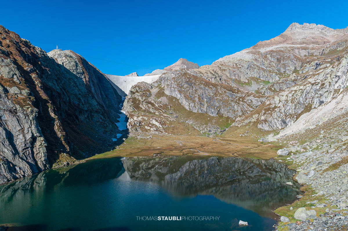 Lago Bianco im Hochgebirge des Tessins, im Hintergrund die Staumauer des Lago dei Cavagnöö in felsiger alpiner Landschaft.