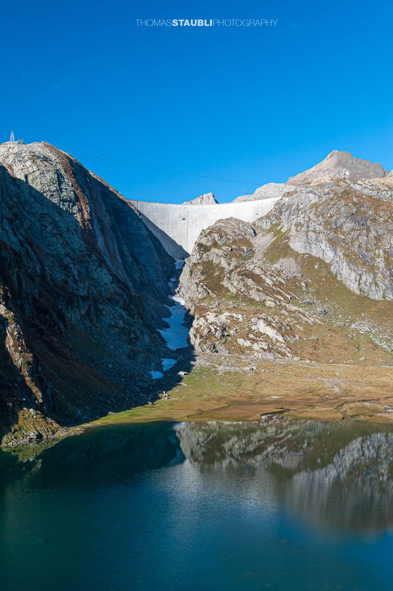 Lago Bianco im Hochgebirge des Tessins, im Hintergrund die Staumauer des Lago dei Cavagnöö in felsiger alpiner Landschaft.