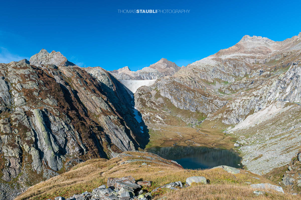 Lago Bianco im Hochgebirge des Tessins, im Hintergrund die Staumauer des Lago dei Cavagnöö in felsiger alpiner Landschaft.