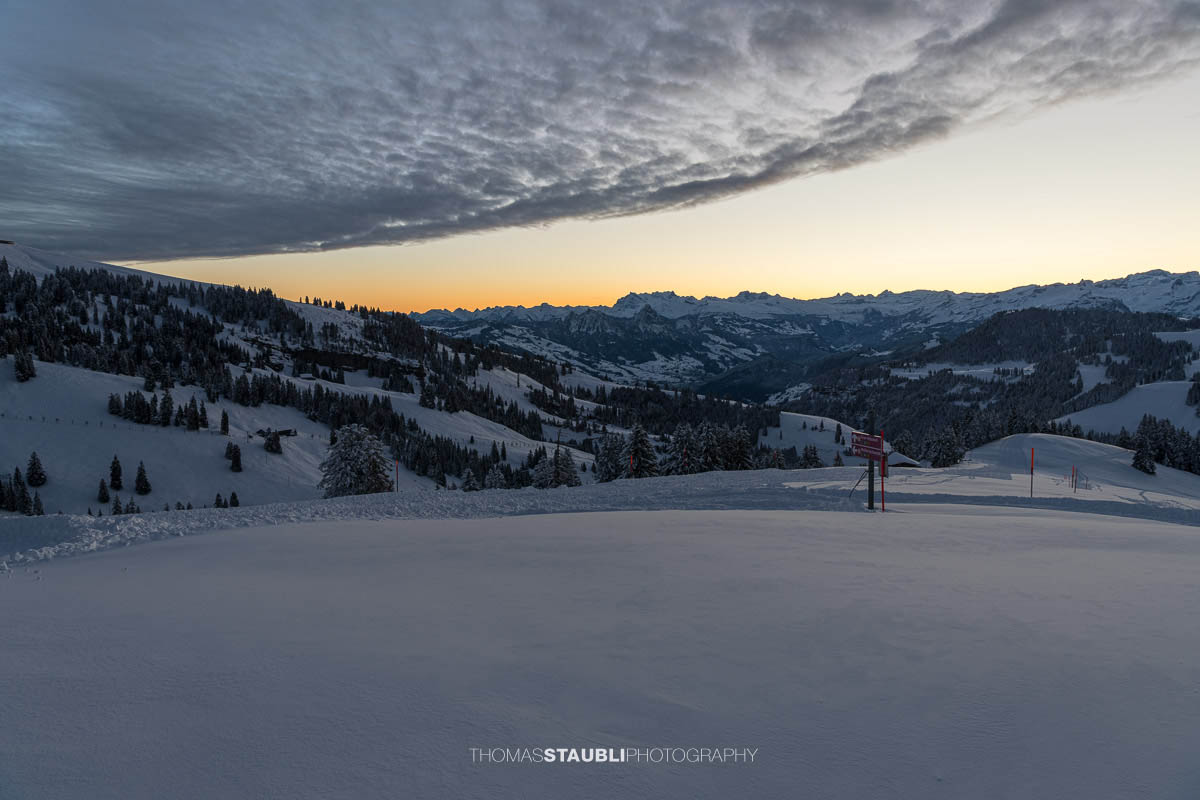 Winterliche Morgendämmerung auf der Rigi: Blick vom Rigi Rotstock zu den Glarner Alpen mit sanften Hängen, verschneiten Wäldern und einem farbigen Horizont.