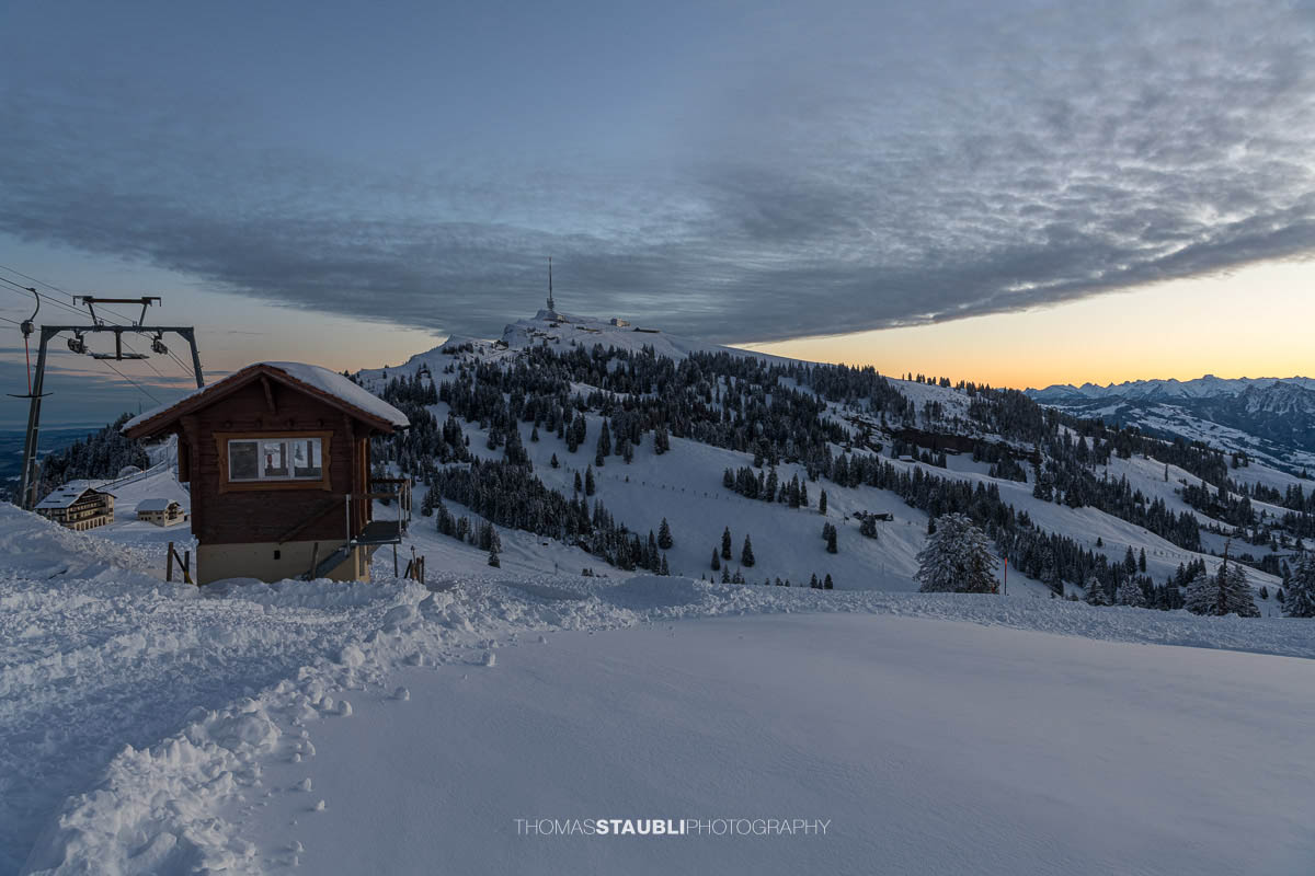 Winterliche Morgendämmerung auf der Rigi: Blick vom Rigi Rotstock zum verschneiten Rigi Kulm, mit sanften Hängen, verschneiten Wäldern und einem farbigen Horizont.