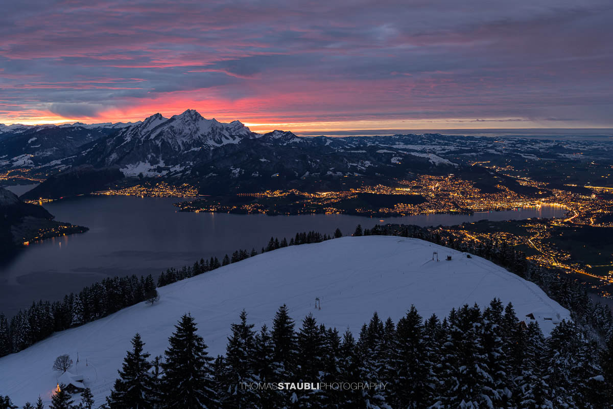 Winterlicher Blick vom Rigi Rotstock auf die beleuchtete Stadt Luzern, den Vierwaldstättersee und den Pilatus im intensiven Abendrot unter einer dramatisch bewölkten Himmeldecke.