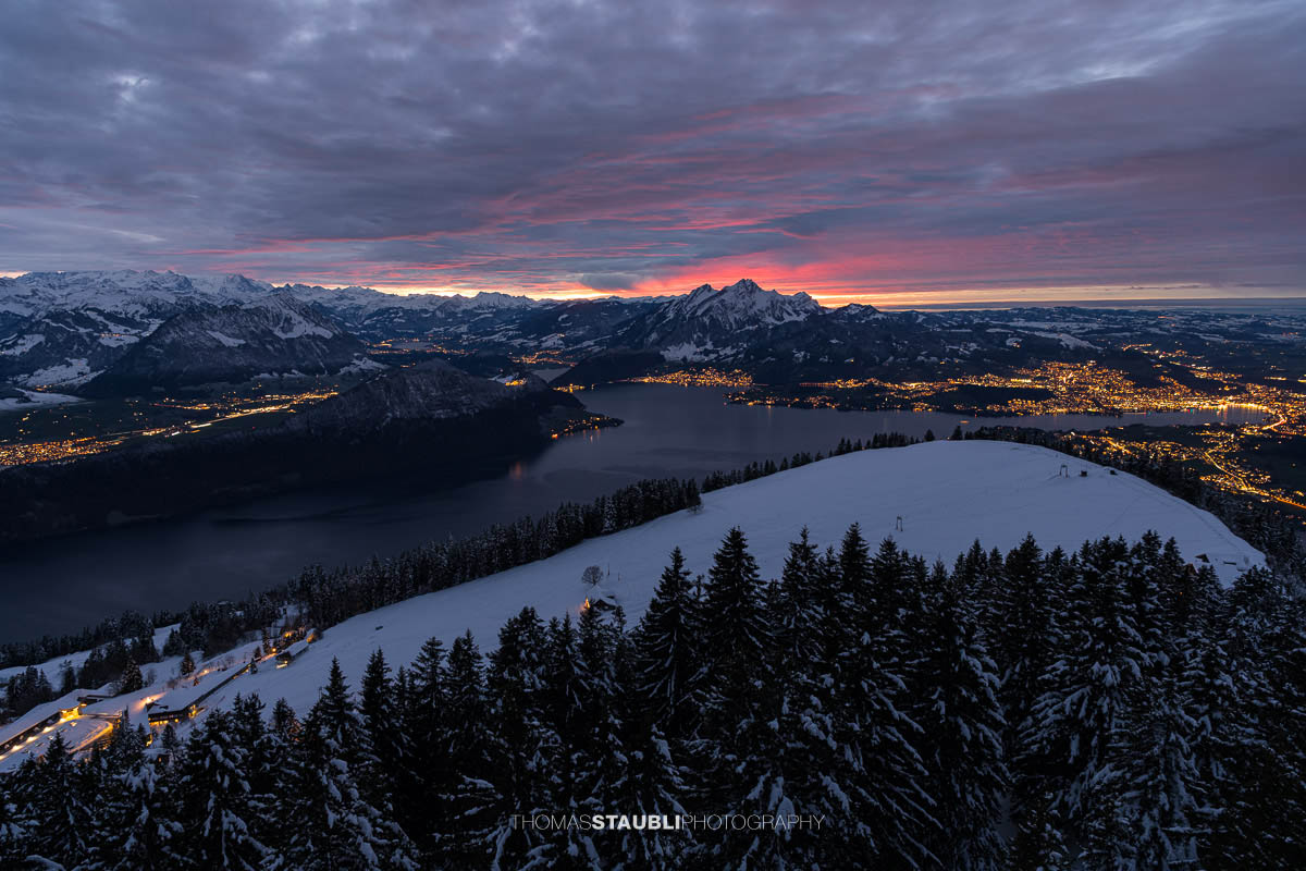 Winterlicher Blick vom Rigi Rotstock auf die beleuchtete Stadt Luzern, den Vierwaldstättersee und den Pilatus im intensiven Abendrot unter einer dramatisch bewölkten Himmeldecke.