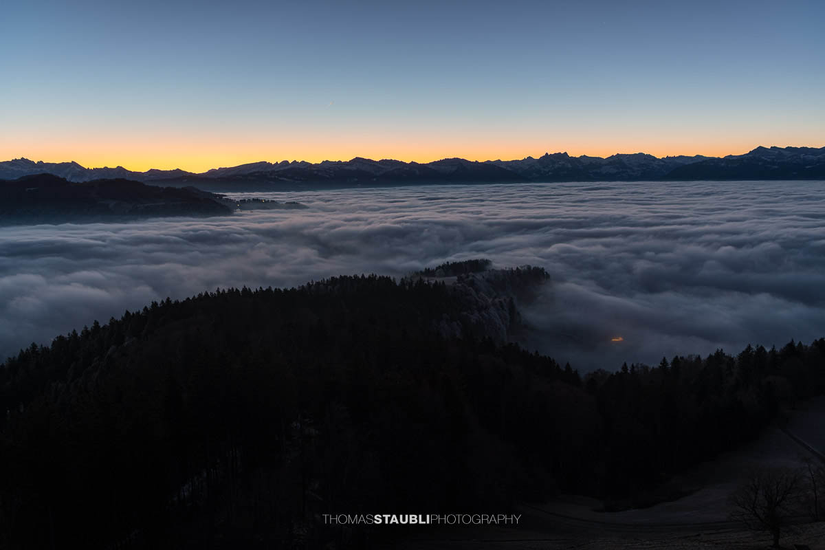 Blick vom Bachtel bei Hinwil in der Morgendämmerung gegen Osten, mit ausgedehntem Nebelmeer über der Ostschweiz und dunklen Hügelzügen im Vordergrund.