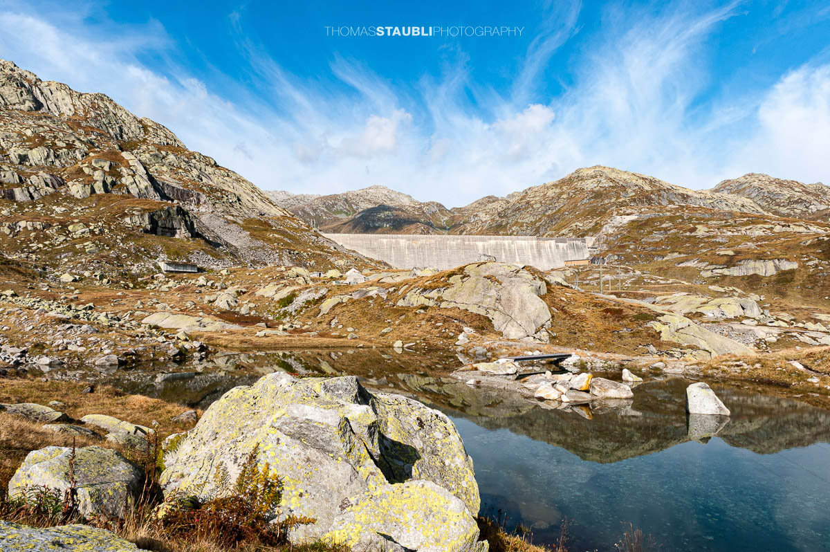 Kleines Bergseelein am Gotthardpass mit klarer Wasseroberfläche und Spiegelung der Felsen, im Hintergrund die Staumauer des Lago di Lucendro.