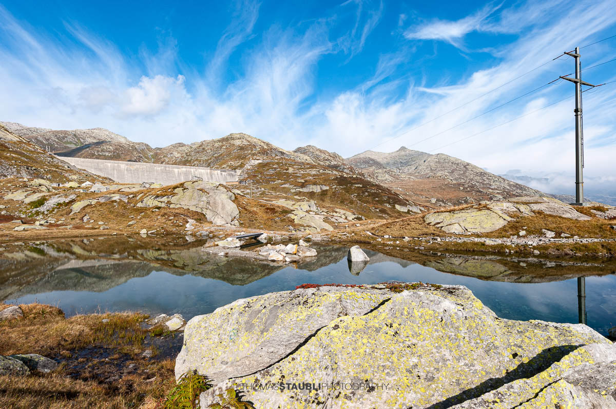 Kleines Bergseelein am Gotthardpass mit klarer Wasseroberfläche und Spiegelung der Felsen, im Hintergrund die Staumauer des Lago di Lucendro.