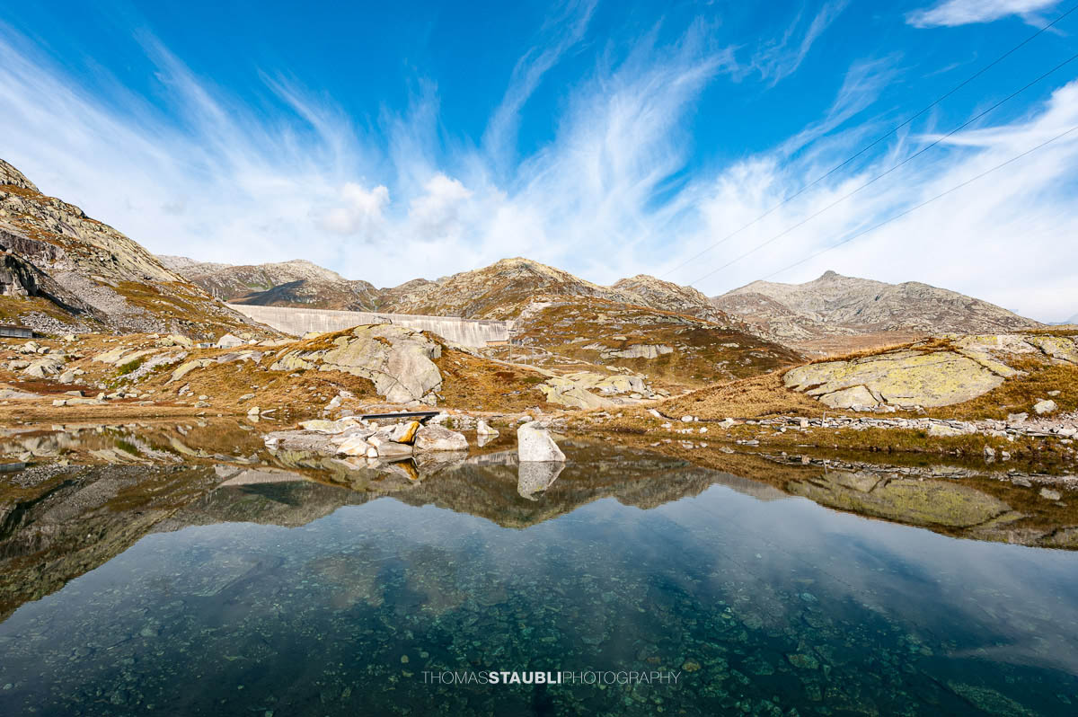 Kleines Bergseelein am Gotthardpass mit klarer Wasseroberfläche und Spiegelung der Felsen, im Hintergrund die Staumauer des Lago di Lucendro.