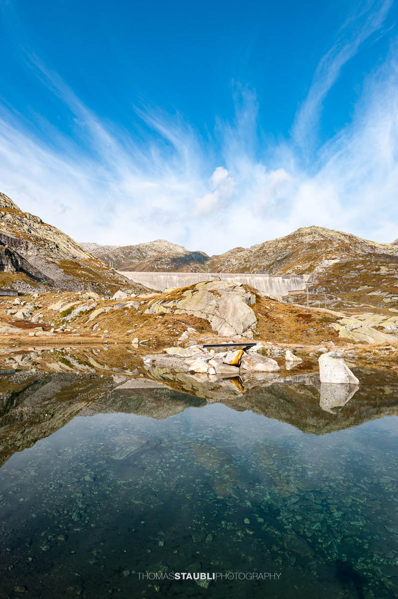 Kleines Bergseelein am Gotthardpass mit klarer Wasseroberfläche und Spiegelung der Felsen, im Hintergrund die Staumauer des Lago di Lucendro.