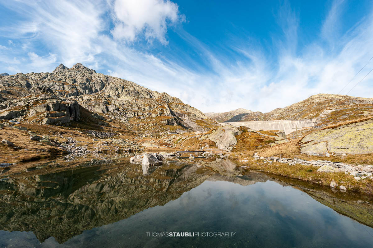 Kleines Bergseelein am Gotthardpass mit klarer Wasseroberfläche und Spiegelung der Felsen, im Hintergrund die Staumauer des Lago di Lucendro.