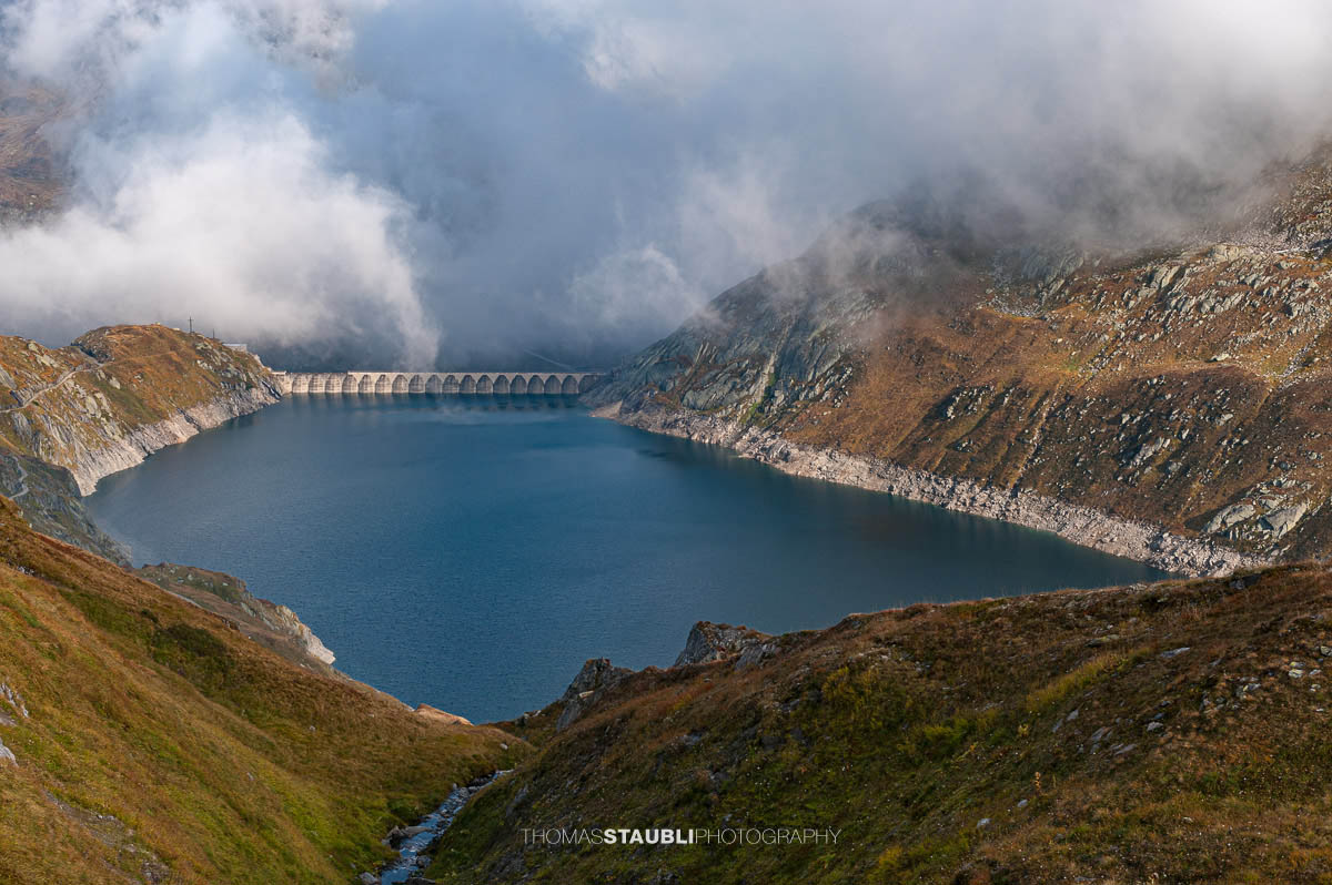 Lago di Lucendro am Gotthardpass, Hochgebirgsstausee mit markanter Staumauer, umgeben von felsigen Hängen und aufziehenden Wolken.