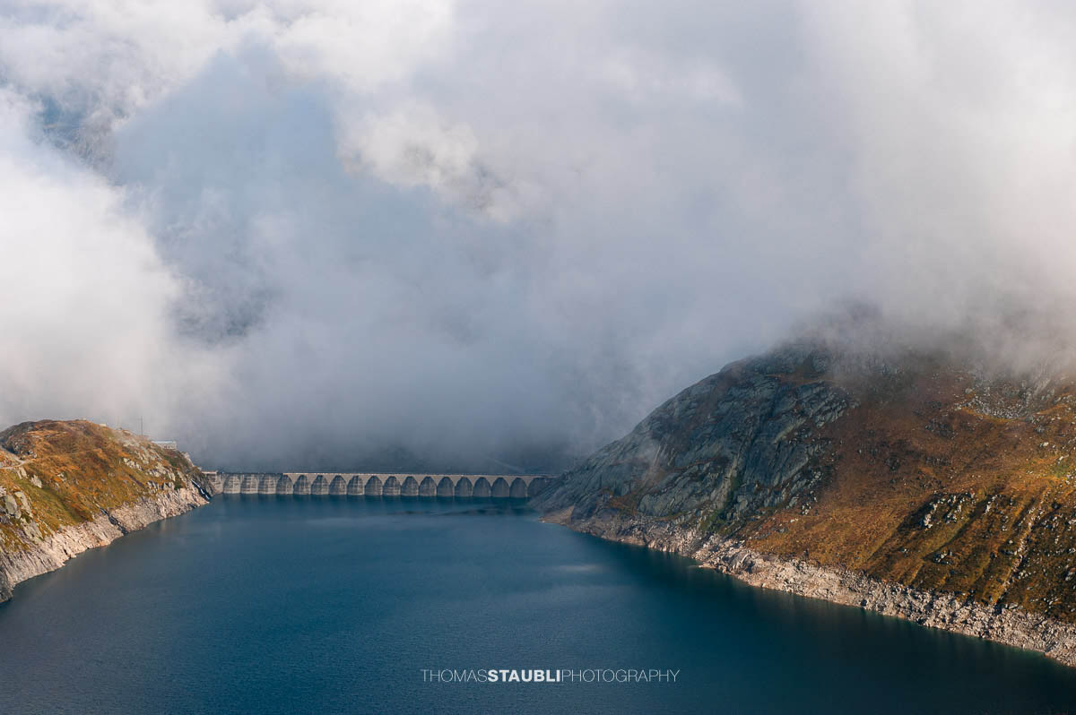 Lago di Lucendro am Gotthardpass, Hochgebirgsstausee mit markanter Staumauer, umgeben von felsigen Hängen und aufziehenden Wolken.