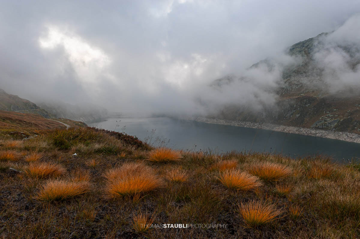 Lago di Lucendro am Gotthardpass mit Staumauer, herbstlicher Vegetation im Vordergrund und aufziehendem Nebel über dem See.