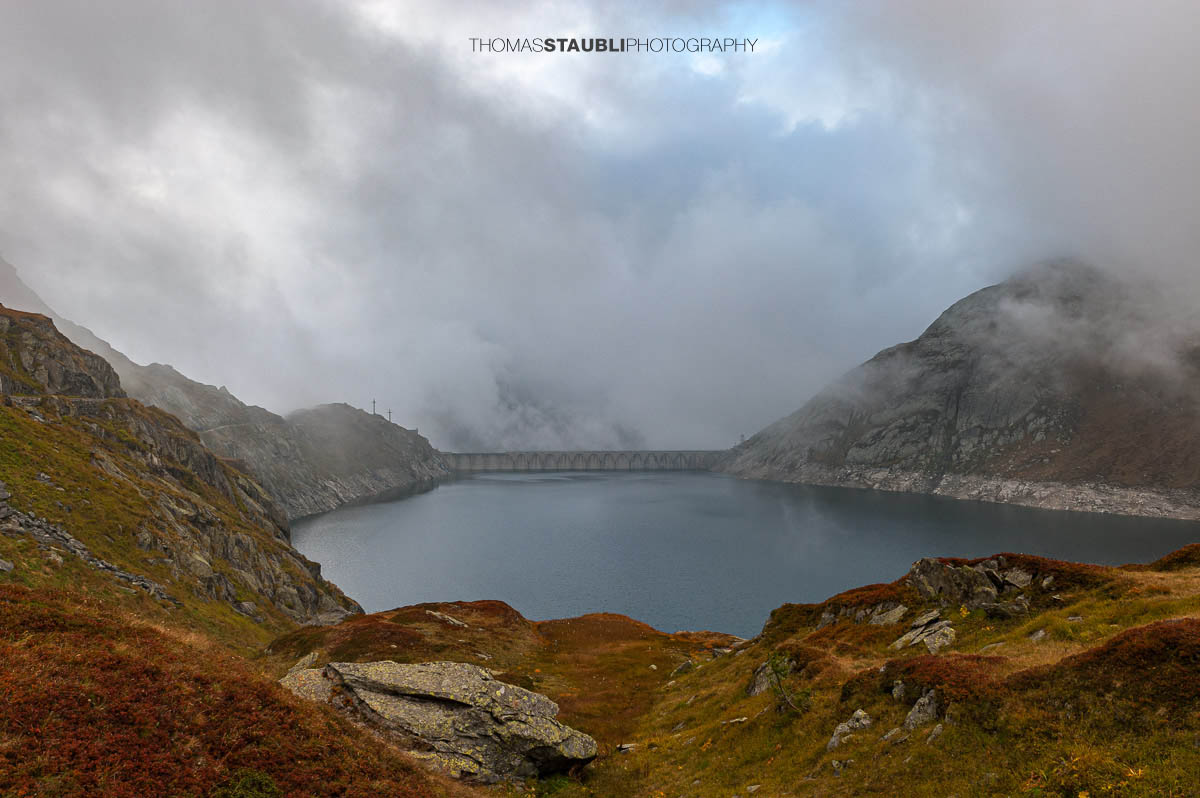 Lago di Lucendro am Gotthardpass, Hochgebirgsstausee mit markanter Staumauer, umgeben von felsigen Hängen und aufziehenden Wolken.
