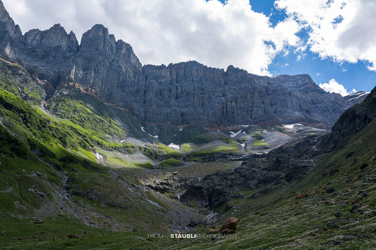 Aufstieg zur Fridolinshütte durch ein steiles alpines Kar mit grünen Matten, Geröllfeldern und einzelnen Schneeresten; darüber aufragende, schroffe Kalkwände im wechselnden Licht.
