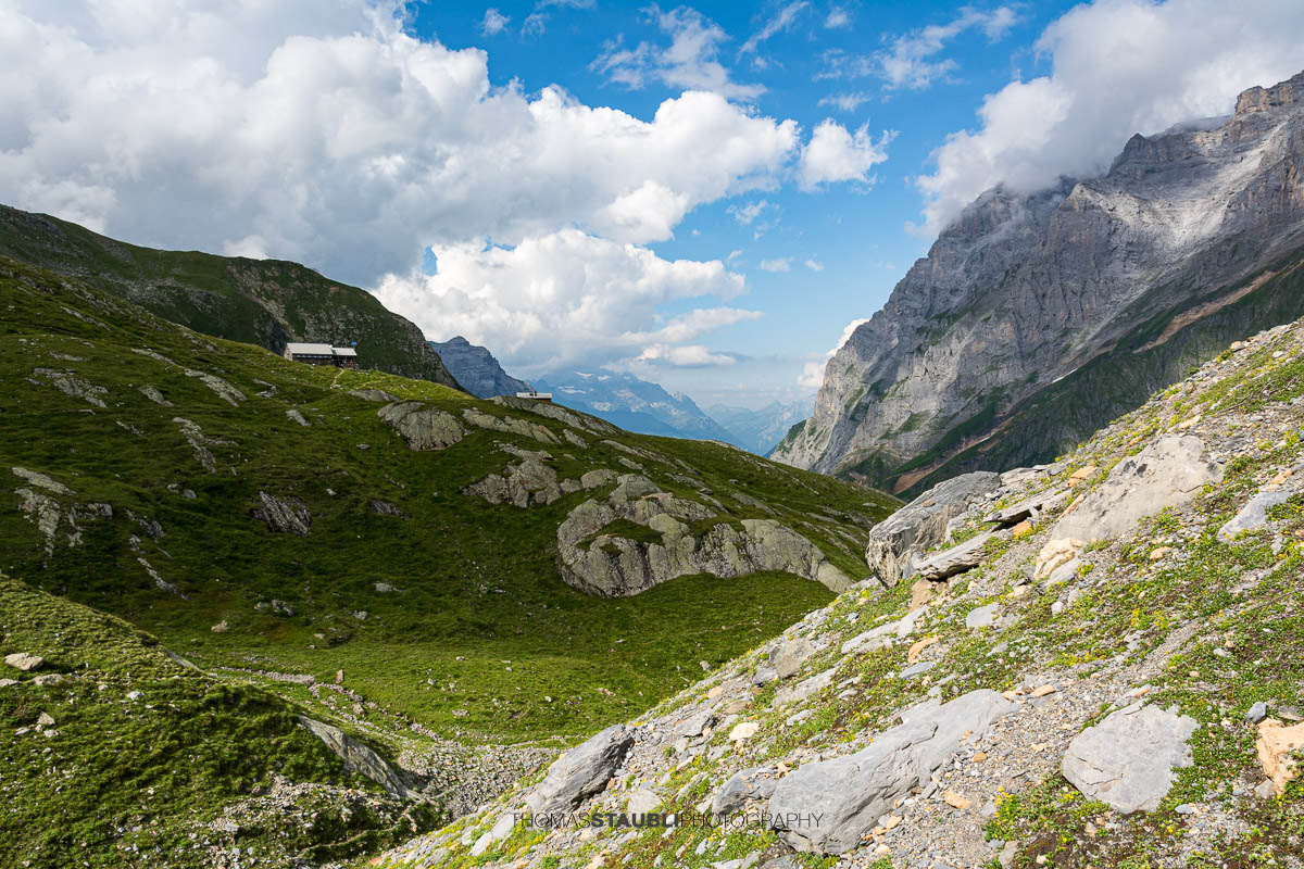 Blick über grüne Alpweiden und felsiges Gelände zur Fridolinshütte, eingebettet zwischen steilen Bergflanken unter einem wechselhaften Sommerhimmel.