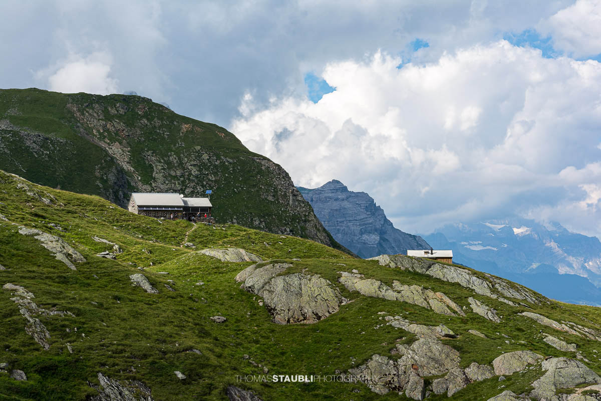 Blick über grüne Alpweiden und felsiges Gelände zur Fridolinshütte, eingebettet zwischen steilen Bergflanken unter einem wechselhaften Sommerhimmel.
