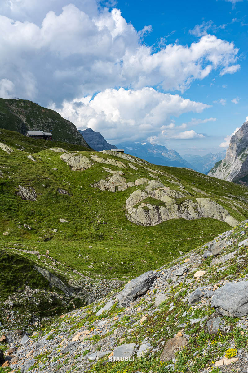 Blick über grüne Alpweiden und felsiges Gelände zur Fridolinshütte, eingebettet zwischen steilen Bergflanken unter einem wechselhaften Sommerhimmel.
