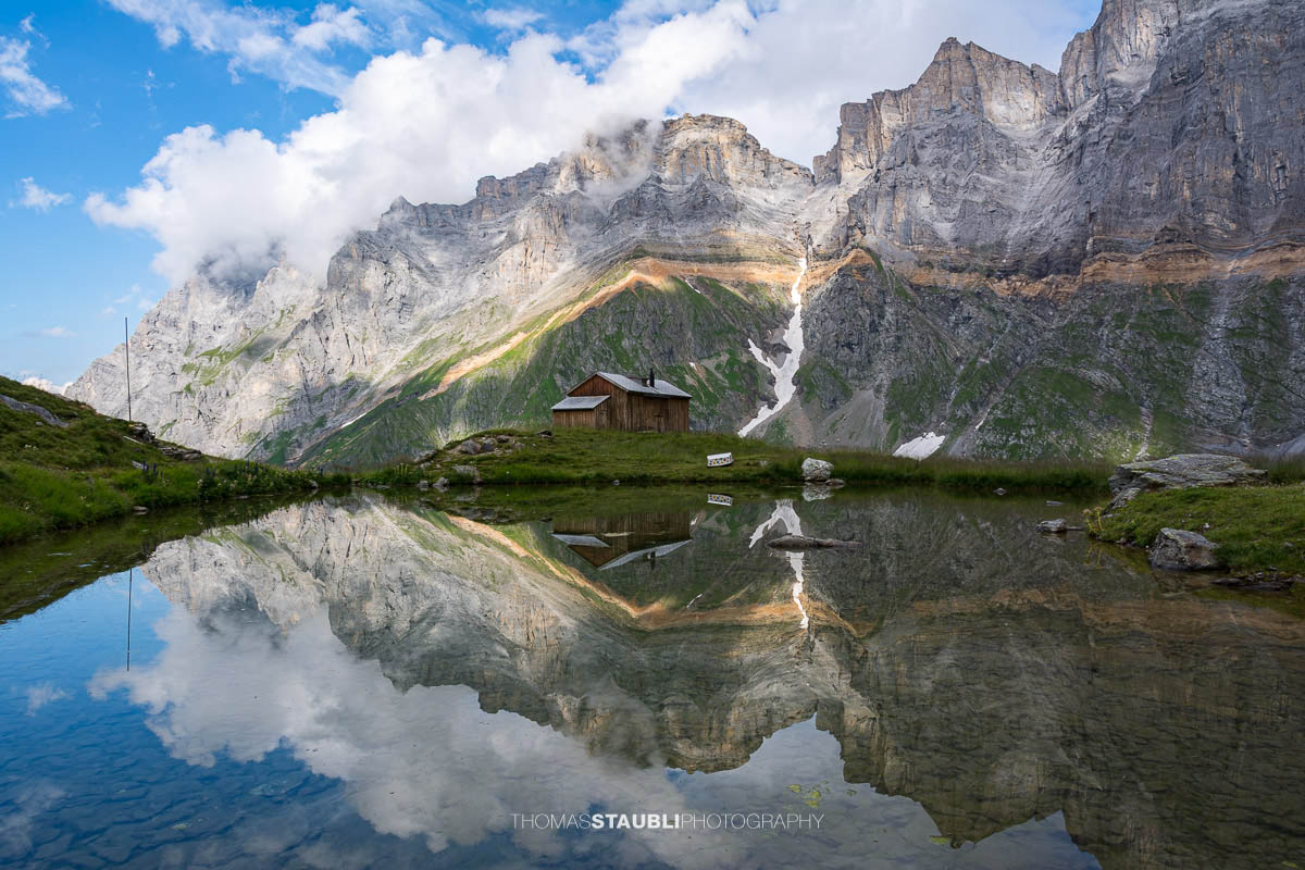 Kleines Bergseelein nahe der Fridolinshütte, in dessen ruhiger Wasserfläche sich die Felswände der Vorder Schiben und der Hinter Schiben spiegeln; alpine Wiesen und ein einzelnes Hüttenhaus im Vordergrund.