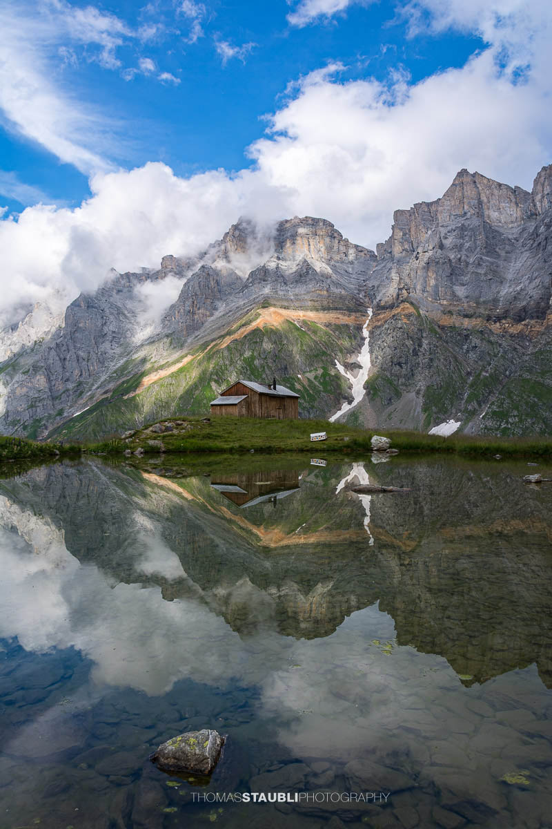 Kleines Bergseelein nahe der Fridolinshütte, in dessen ruhiger Wasserfläche sich die Felswände der Vorder Schiben und der Hinter Schiben spiegeln; alpine Wiesen und ein einzelnes Hüttenhaus im Vordergrund.