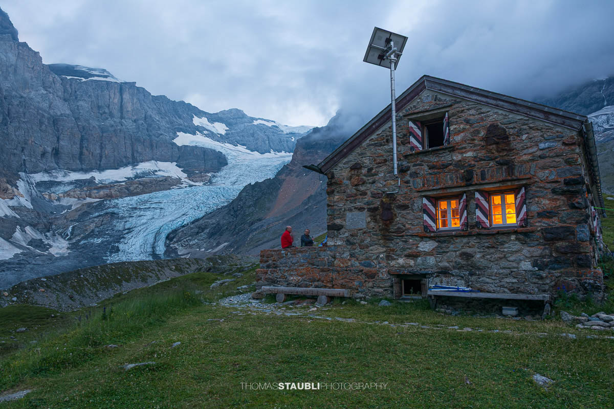 Fridolinshütte im Kanton Glarus in der Abenddämmerung, dahinter der Gletscher Bifertenfirn unter steilen Felswänden.