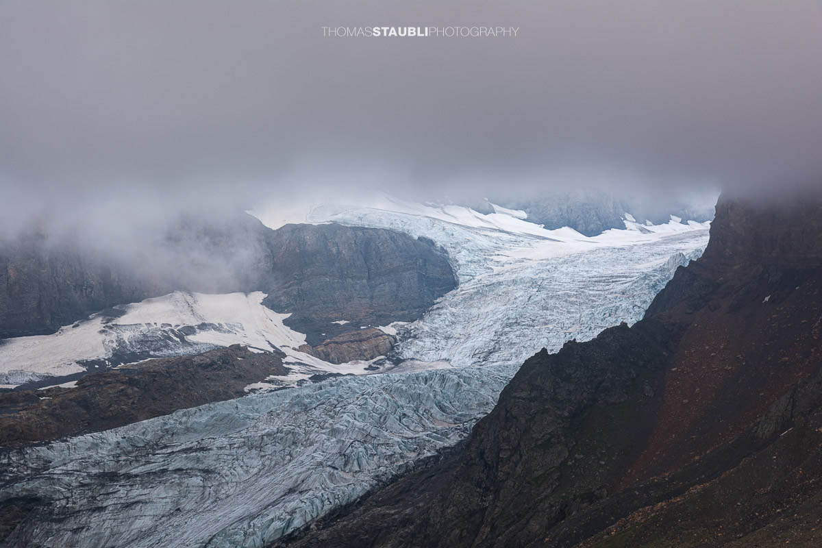 Blick auf den Bifertenfirn im Jahr 2013 im Kanton Glarus, teilweise von tief hängenden Wolken umgeben, mit sichtbarem Gletscherstrom, Schuttflächen und steilen Felswänden in alpiner Hochgebirgslandschaft.