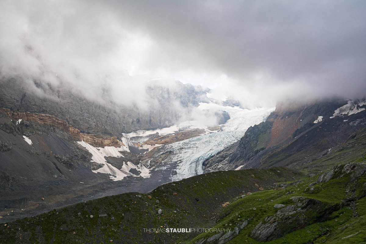 Blick auf den Bifertenfirn im Jahr 2013 im Kanton Glarus, teilweise von tief hängenden Wolken umgeben, mit sichtbarem Gletscherstrom, Schuttflächen und steilen Felswänden in alpiner Hochgebirgslandschaft.