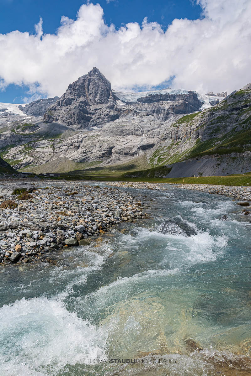 glasklares Wasser des Oberstafelbachs auf der Alp Ober Sand im Kanton Glarus; im Hintergrund in der Spitzalpelistock, rechts davon der Claridenfirn, umgeben von Geröllflächen, alpiner Ebene und steilen Felswänden.