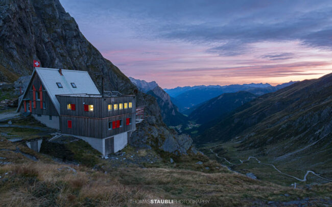 Die Capanna Scaletta liegt in der Abenddämmerung auf einem felsigen Hang oberhalb des Bleniotals, umgeben von alpiner Landschaft und einem weit geöffneten Bergtal.