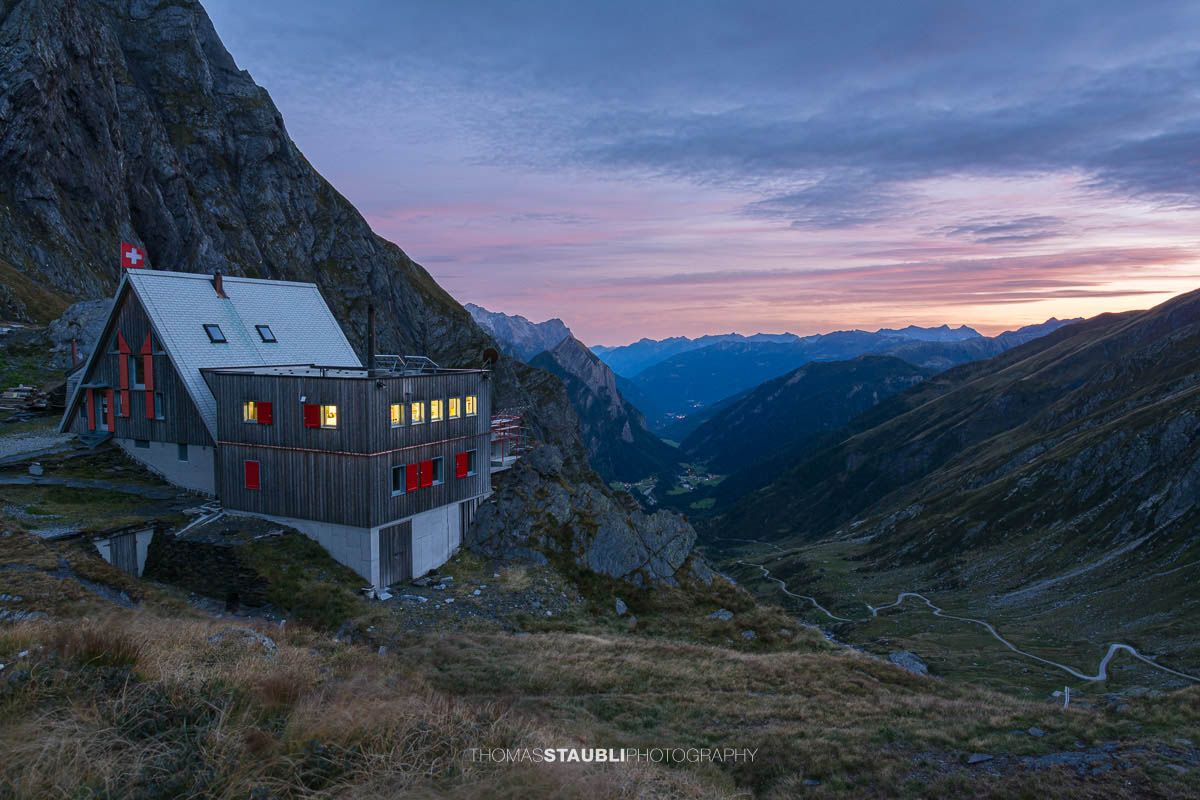 Die Capanna Scaletta liegt in der Abenddämmerung auf einem felsigen Hang oberhalb des Bleniotals, umgeben von alpiner Landschaft und einem weit geöffneten Bergtal.