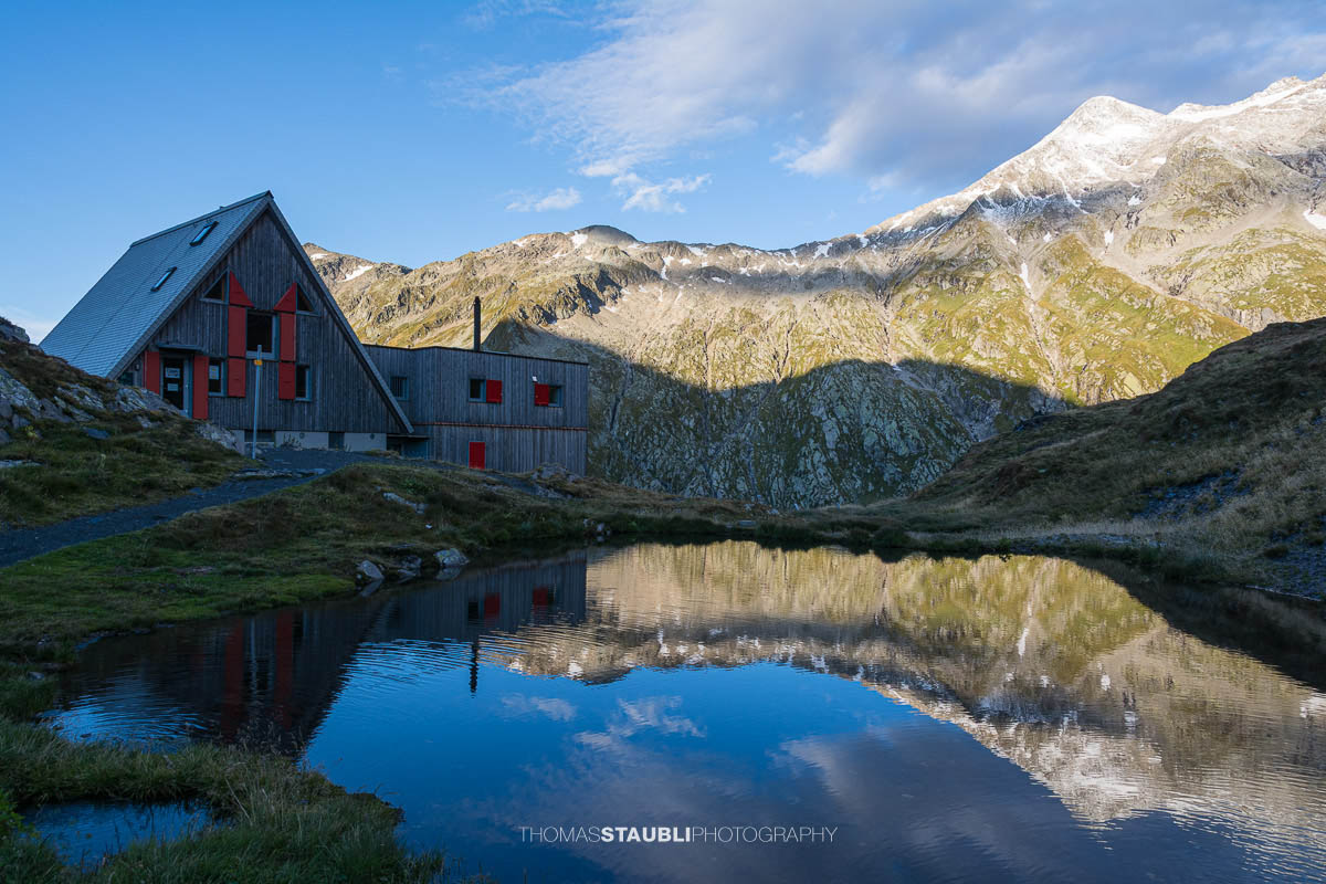 Die Capanna Scaletta im Tessin spiegelt sich in einem kleinen Bergseelein, umgeben von alpinen Hängen und felsigen Gipfeln der Greinaregion.