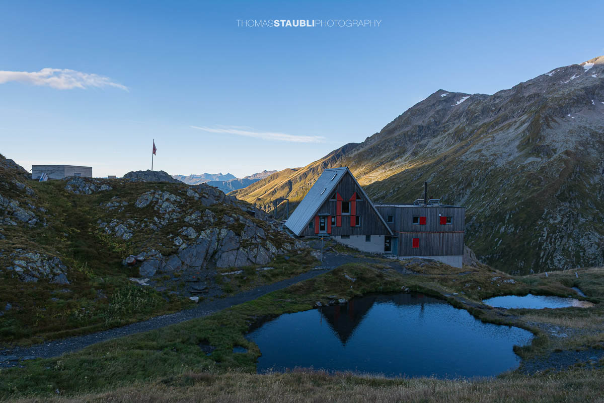 Die Capanna Scaletta im Tessin spiegelt sich in einem kleinen Bergseelein, umgeben von alpinen Hängen und felsigen Gipfeln der Greinaregion.