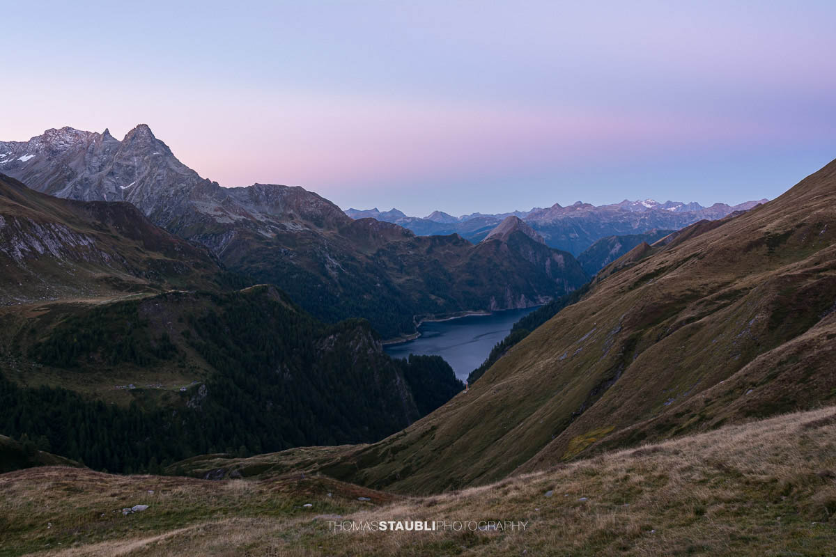 Morgendämmerung oberhalb der Capanna Motterascio mit Blick über alpine Hänge zum Lago di Luzzone und den umliegenden Bergketten der Greina-Region im Tessin.