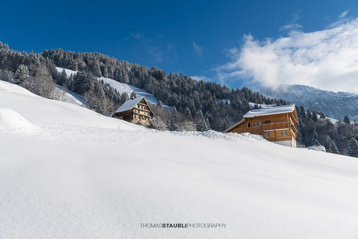 Verschneite Bauernhäuser am Hang über dem Wägitalersee, umgeben von Wald und alpiner Winterlandschaft im Wägital, Kanton Schwyz.