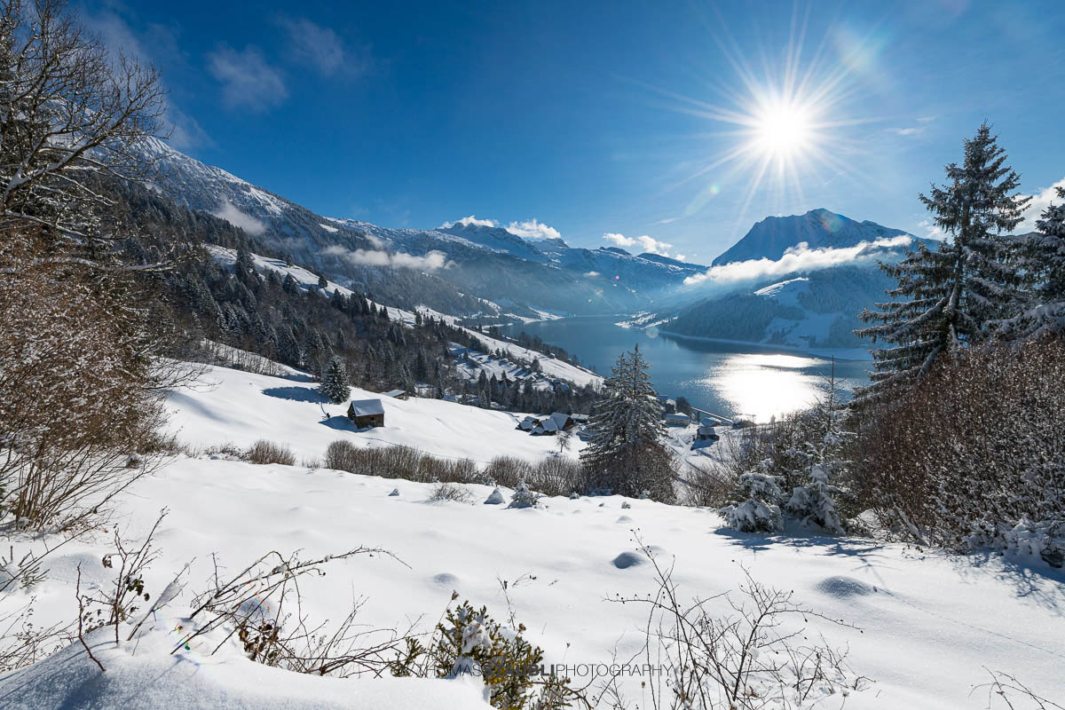 Winterlandschaft mit verschneiten Hängen und Blick auf den Wägitalersee im Wägital, Sonnenstand über dem Tal und umliegende Berge im Kanton Schwyz.