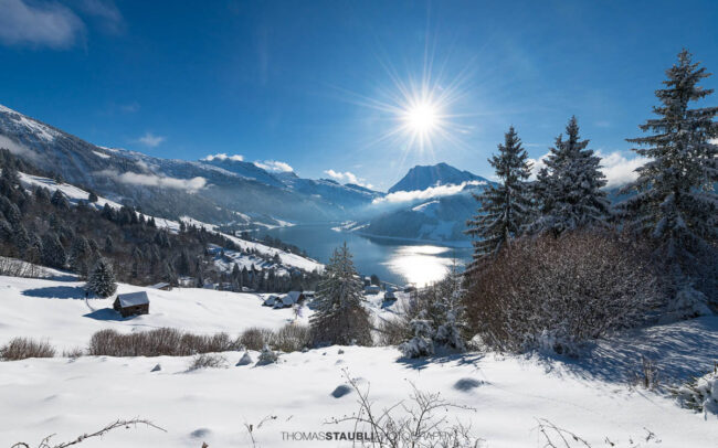 Winterlandschaft mit verschneiten Hängen und Blick auf den Wägitalersee im Wägital, Sonnenstand über dem Tal und umliegende Berge im Kanton Schwyz.