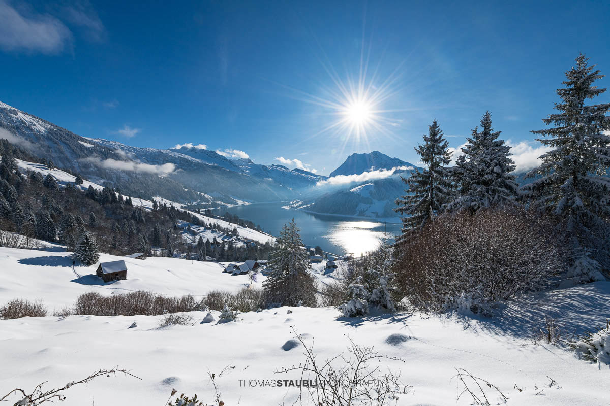 Winterlandschaft mit verschneiten Hängen und Blick auf den Wägitalersee im Wägital, Sonnenstand über dem Tal und umliegende Berge im Kanton Schwyz.