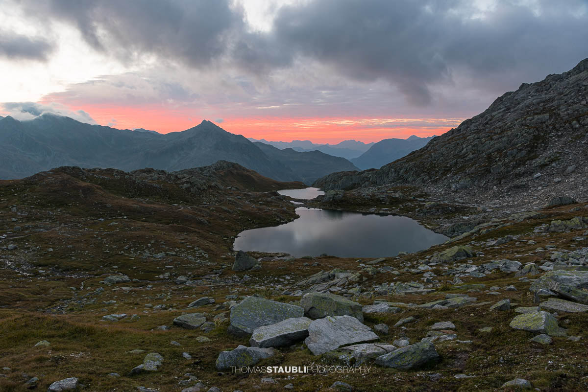 Morgenrot über den Laghi della Valletta: kleine Hochgebirgsseen in felsiger Landschaft, dunkle Wolken und roter Horizont über den Tessiner Alpen.