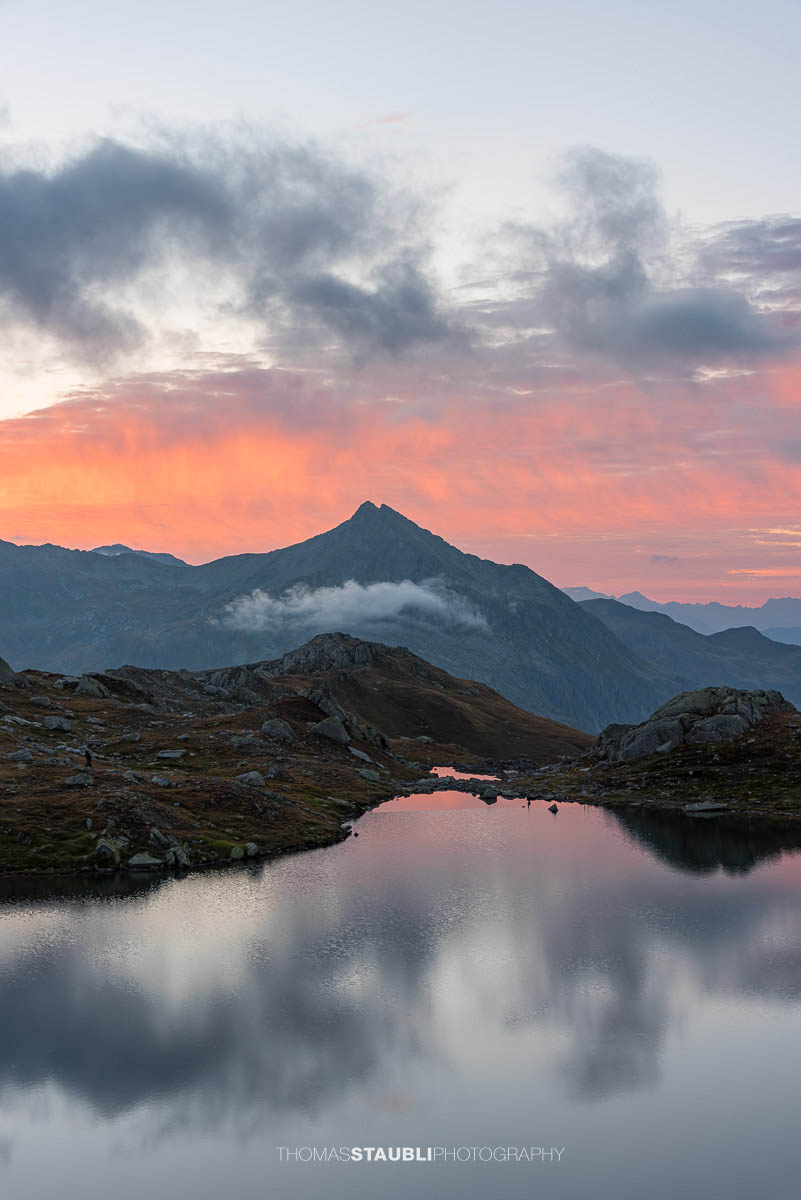 Morgenrot über den Laghi della Valletta: kleine Hochgebirgsseen in felsiger Landschaft, dunkle Wolken und roter Horizont spiegeln sich im ruhigen Wasser der Tessiner Alpen.