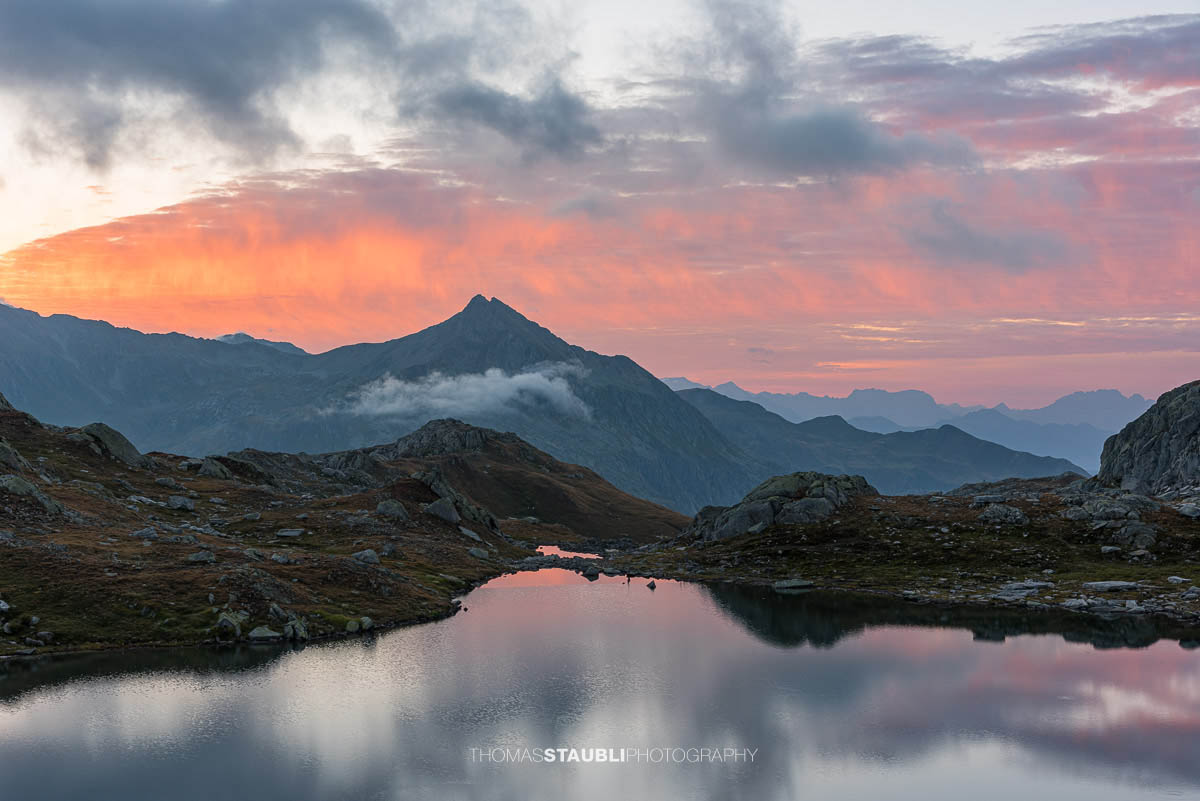 Morgenrot über den Laghi della Valletta: kleine Hochgebirgsseen in felsiger Landschaft, dunkle Wolken und roter Horizont spiegeln sich im ruhigen Wasser der Tessiner Alpen.